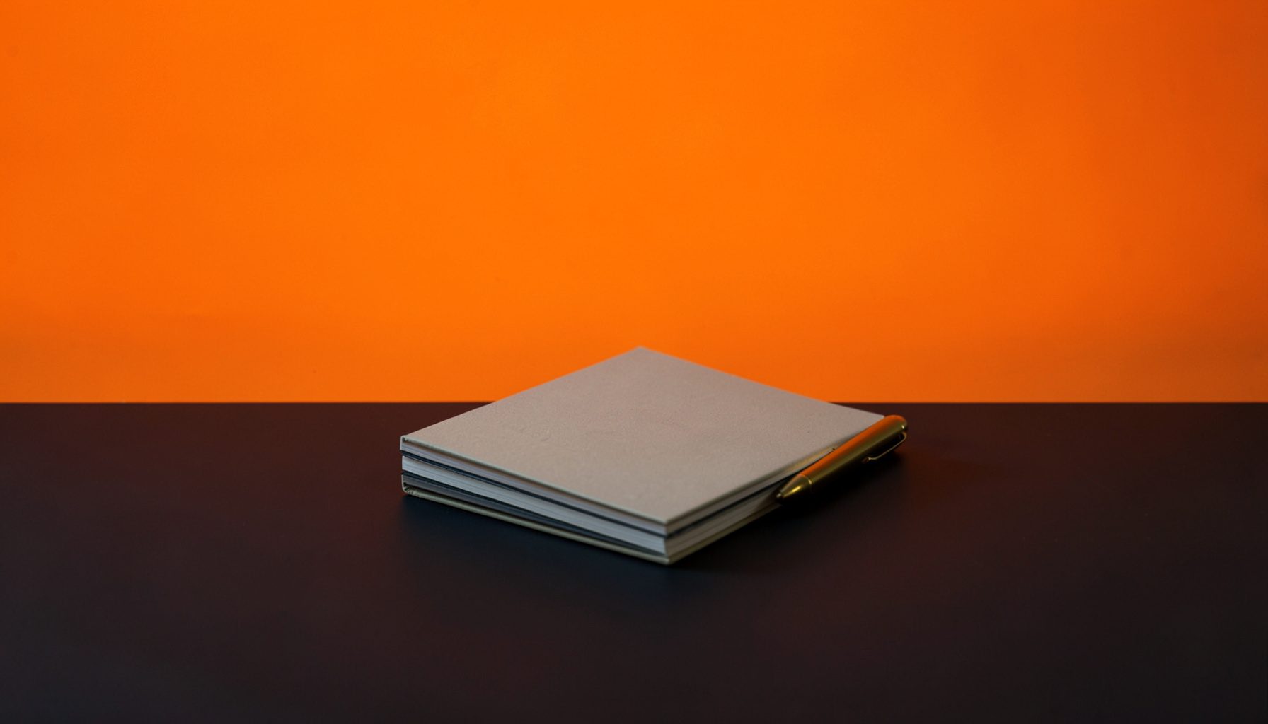 A small stack of neutral-toned notebooks and a pen resting on a dark seamless surface, muted gold accent lighting, minimal and editorial in feel